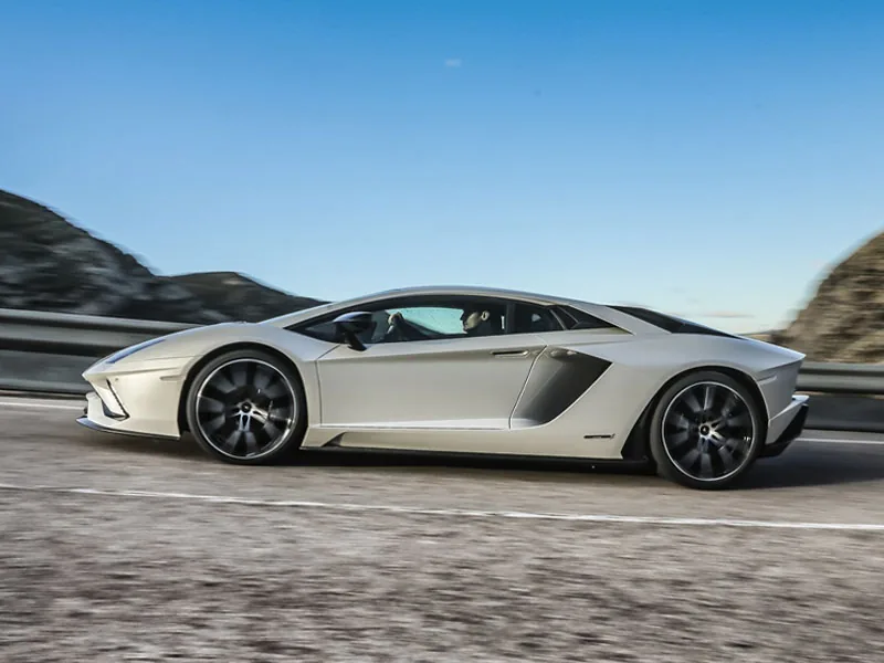 A silver Lamborghini Aventador in side profile on a coast road